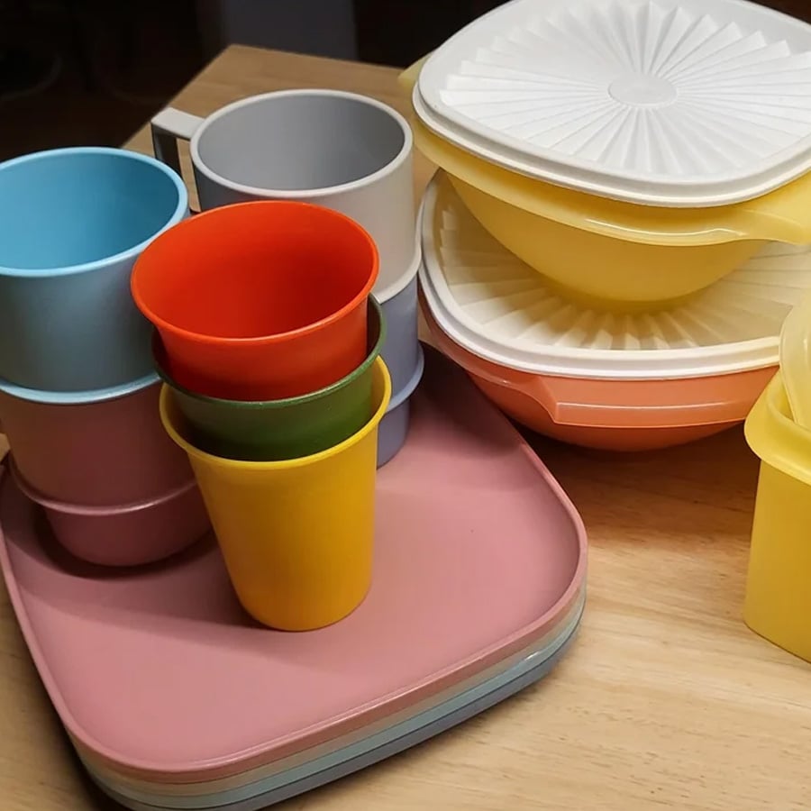 Colorful plastic cups, plates, and bowls are stacked neatly on a wooden table. The items are in pastel and bright colors, with some covered by white lids.