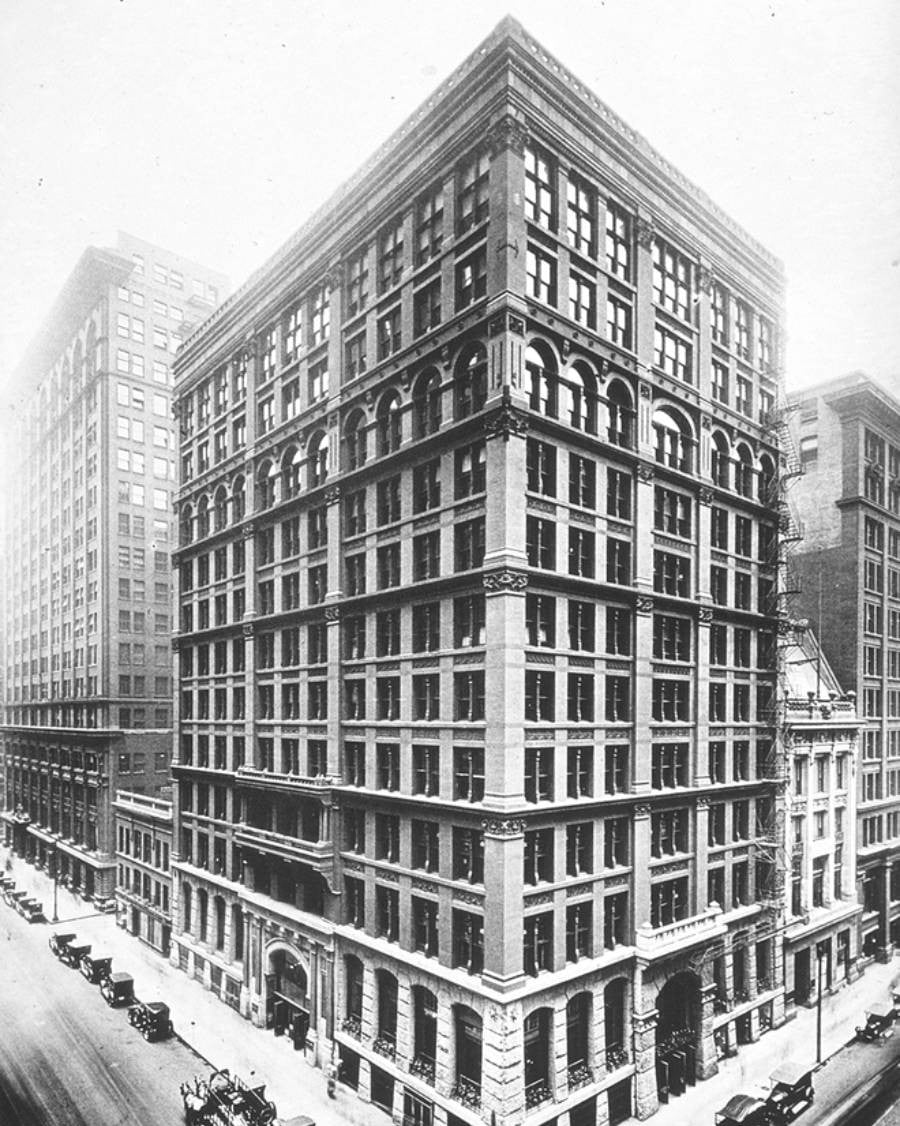 Black-and-white photo of a tall, early skyscraper on a city corner with large windows, arched details, and horse-drawn carriages and early cars lining the surrounding streets.