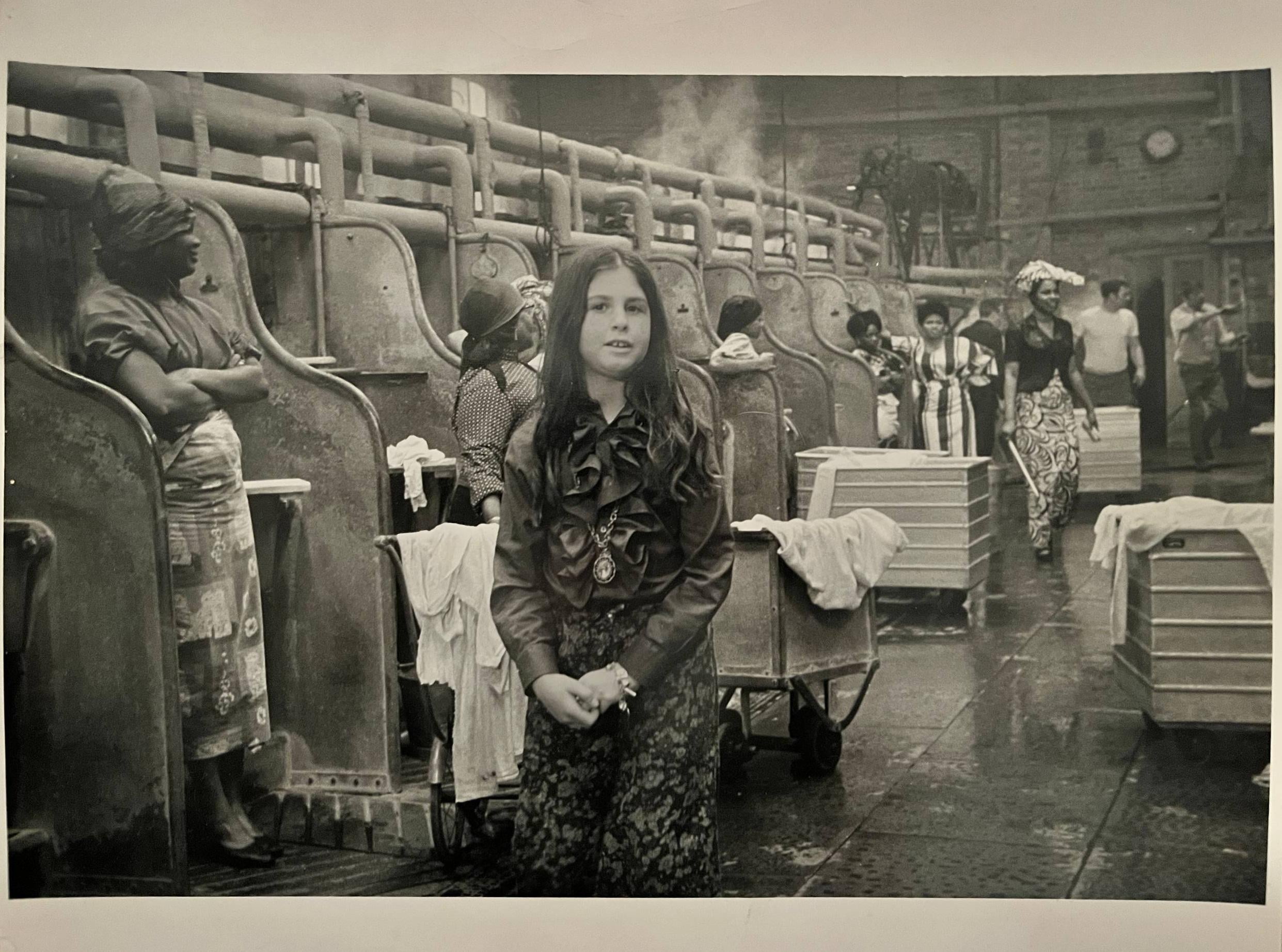 A young girl stands in the foreground of a busy laundry facility, surrounded by women working at large industrial washing stations, with steam rising in the background.