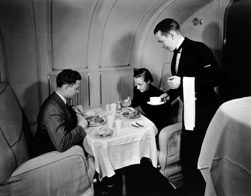 A man and woman dine at a table on an airplane while a waiter in a tuxedo serves them. The scene looks elegant and vintage, likely from the mid-20th century.
