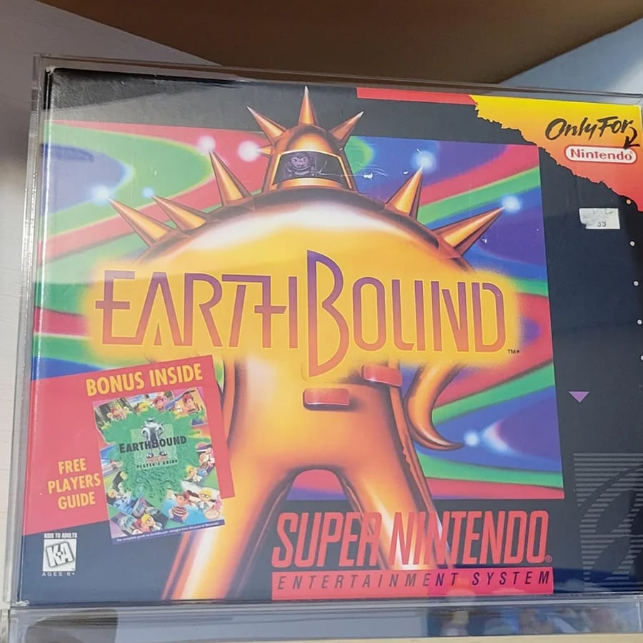 EarthBound Super Nintendo game box art featuring a golden figure with spikes, colorful rays in the background, and a sticker advertising a bonus player’s guide inside.