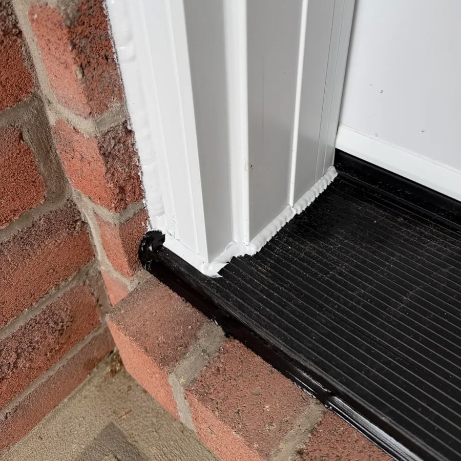 Close-up of a door threshold with white trim, black sealant applied at the base, and red brick on the left side. The black sealant creates a waterproof barrier between the door frame and the ground.