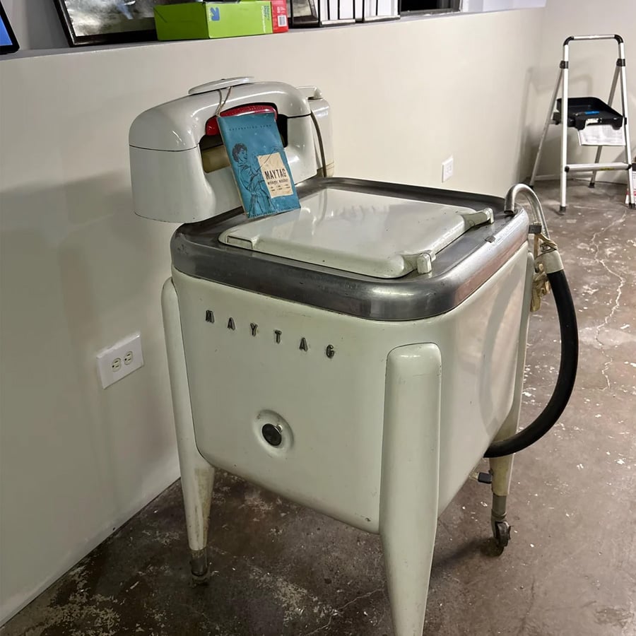 Vintage Maytag washing machine with a wringer on top, standing on a concrete floor near a wall with power outlets. A book rests on the wringer, and a black hose hangs from the side.
