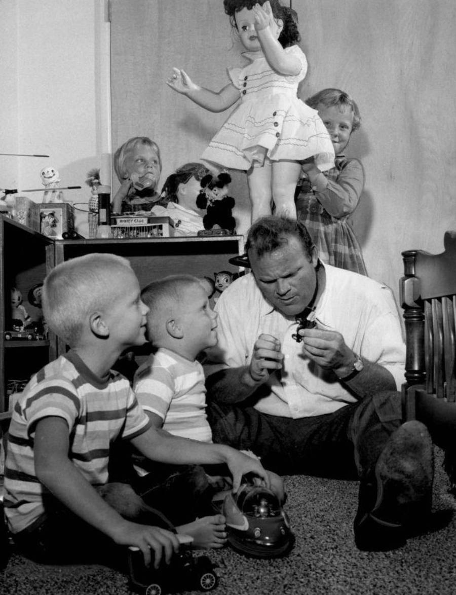 A man sits on the floor playing with two young boys and toy cars, while a woman and two girls, one standing on a table with a doll, look on in a room filled with toys.