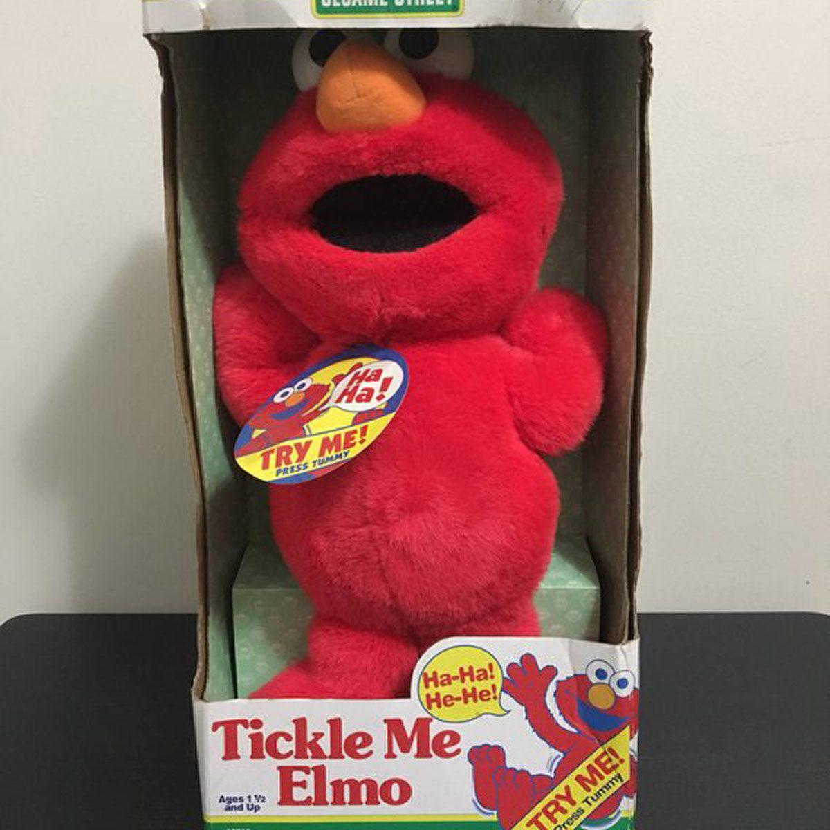 A boxed Tickle Me Elmo plush toy from Sesame Street, with a sticker saying “Ha-Ha! Try Me! Press Here!” and Elmo’s mouth open as if laughing. The box is white with red text and Elmo’s picture.