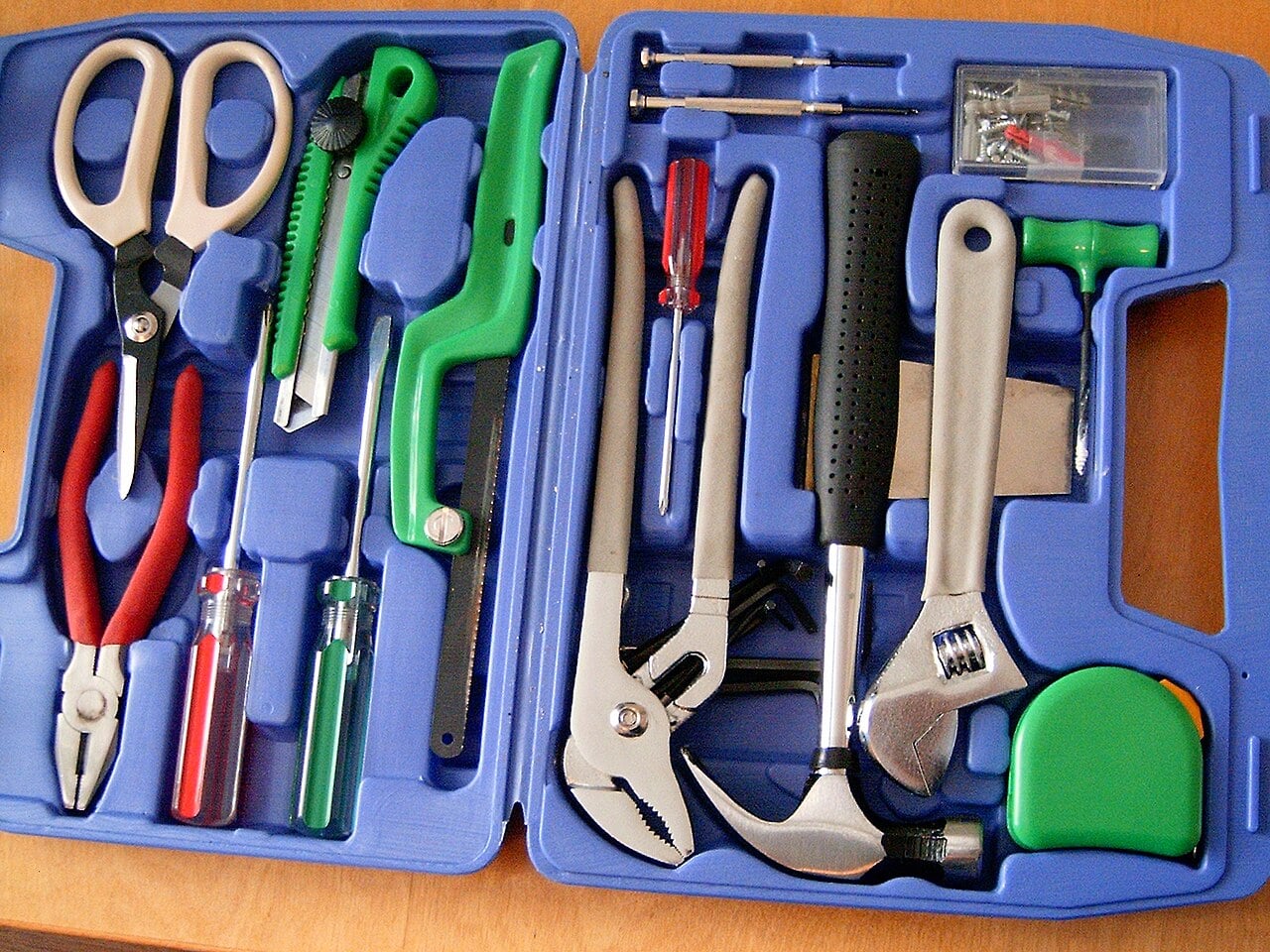A blue tool kit containing scissors, pliers, screwdrivers, a wrench, a hammer, a tape measure, a box cutter, a saw, and other small hand tools, all organized in molded compartments.