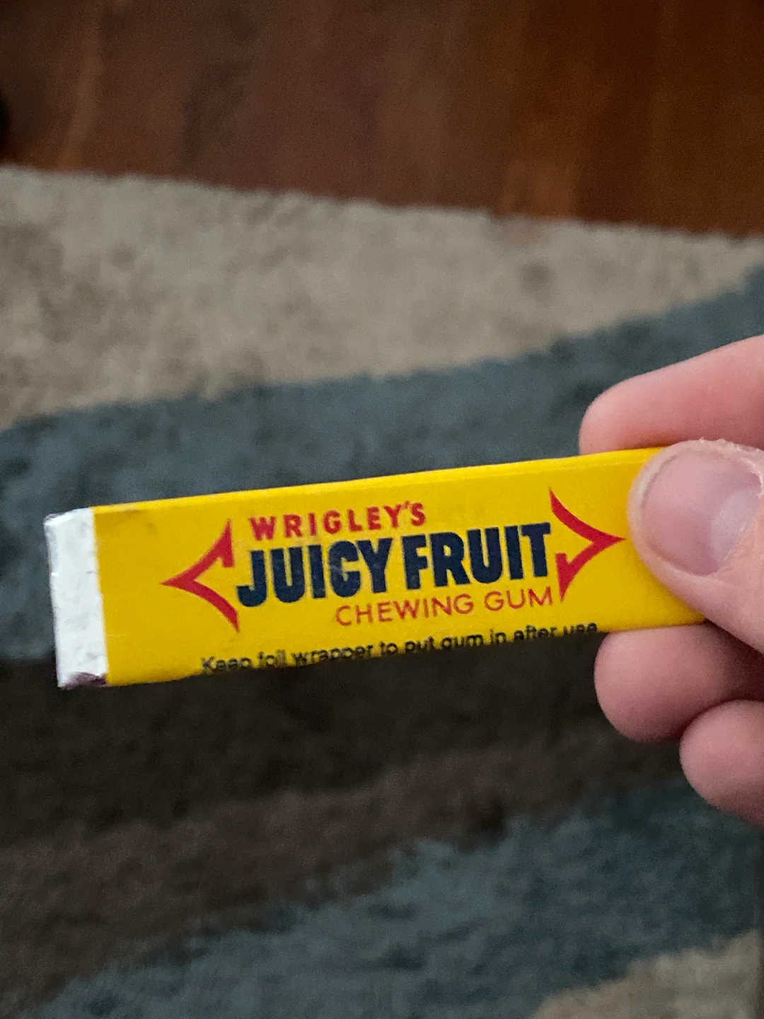 A hand holds a stick of Wrigley’s Juicy Fruit chewing gum with a yellow wrapper, partially unwrapped, against a blurry background of carpet and wood floor.