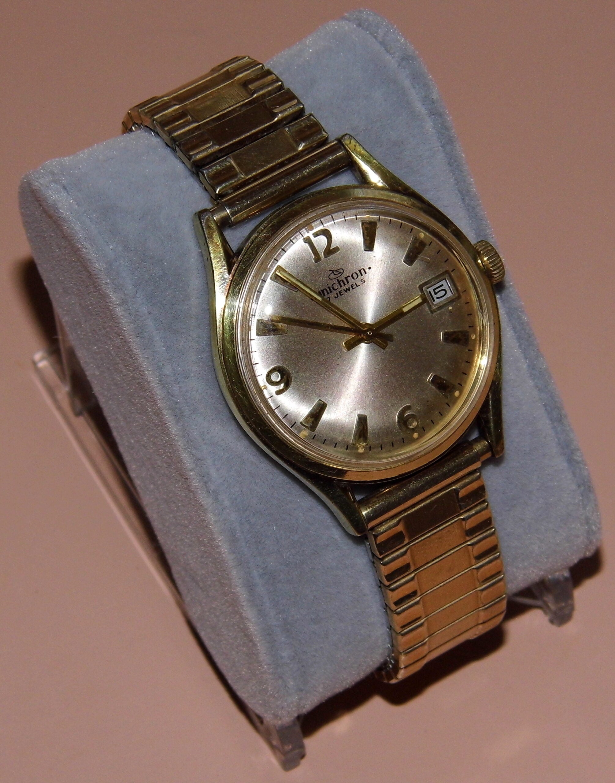 A vintage gold wristwatch with a metal band, silver dial, and numeric markers is displayed on a blue velvet cushion against a plain background.