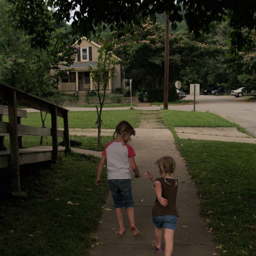 Two young girls walk hand in hand on a sidewalk toward a house, surrounded by greenery and trees. The street is quiet, and the atmosphere is calm and peaceful.