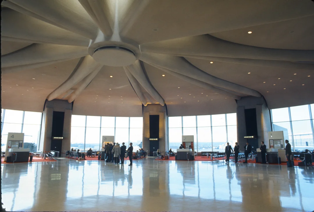 A spacious airport terminal with a high, dome-like ceiling, large windows letting in natural light, and groups of people near gates and seating areas.