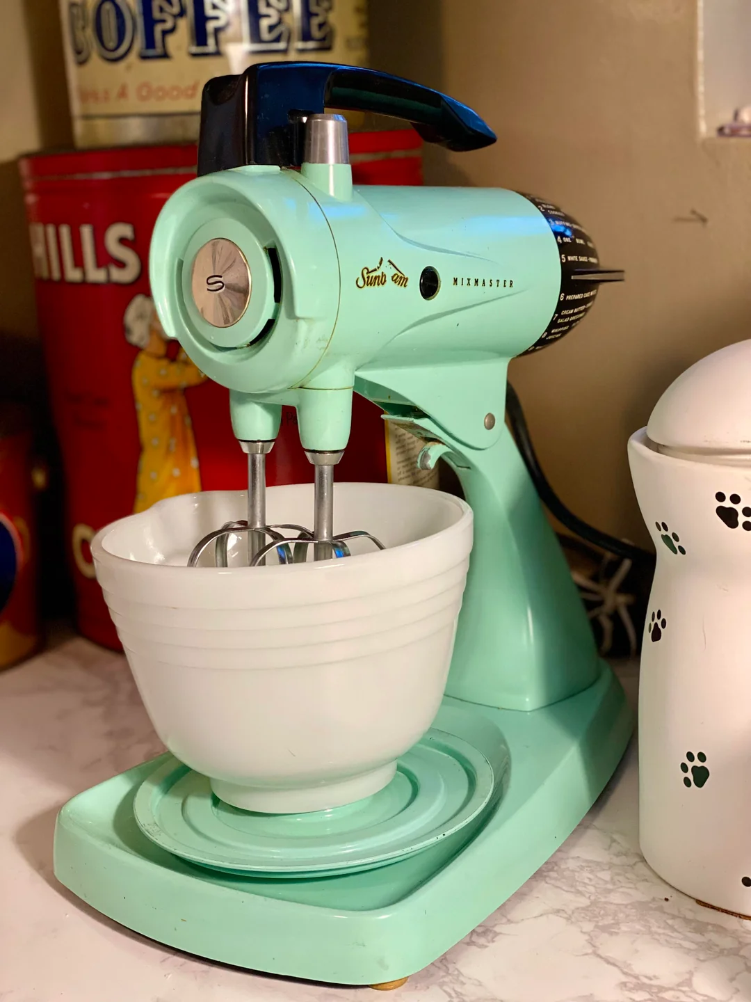 A vintage aqua Sunbeam Mixmaster stand mixer with a white mixing bowl sits on a kitchen counter. In the background are a red Hills Coffee tin and a white container with black paw prints.