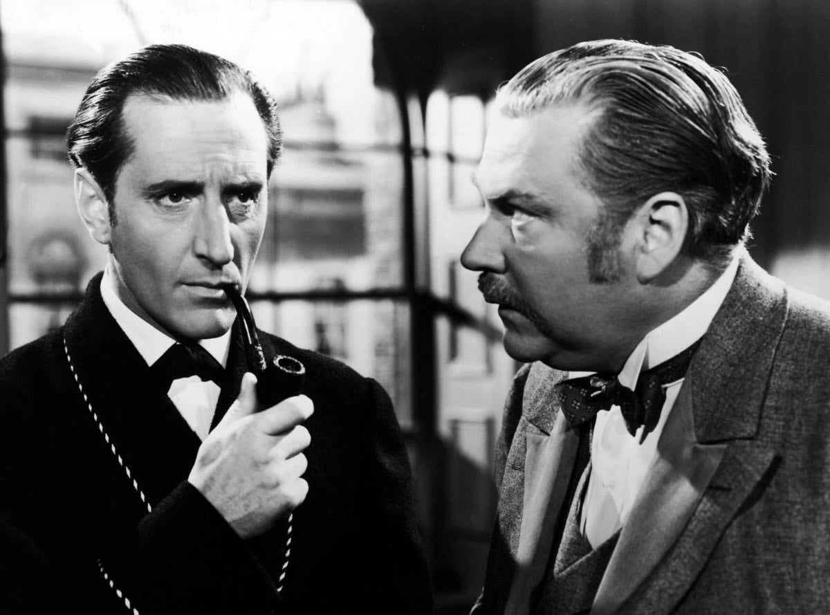 Two men in vintage attire, one holding a pipe thoughtfully while the other listens intently. The background suggests a classic, old-fashioned setting, possibly from an early film or period drama.
