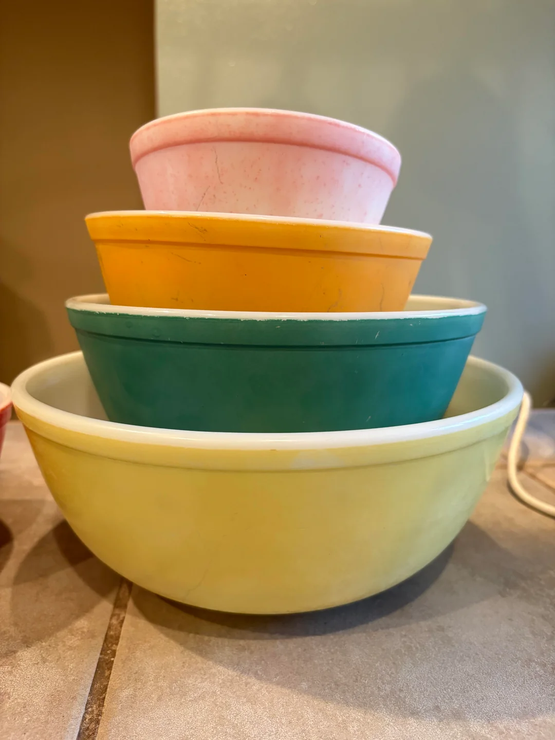 Four vintage mixing bowls in pastel colors—pink, orange, green, and yellow—are stacked inside each other on a tiled countertop, with a soft background.