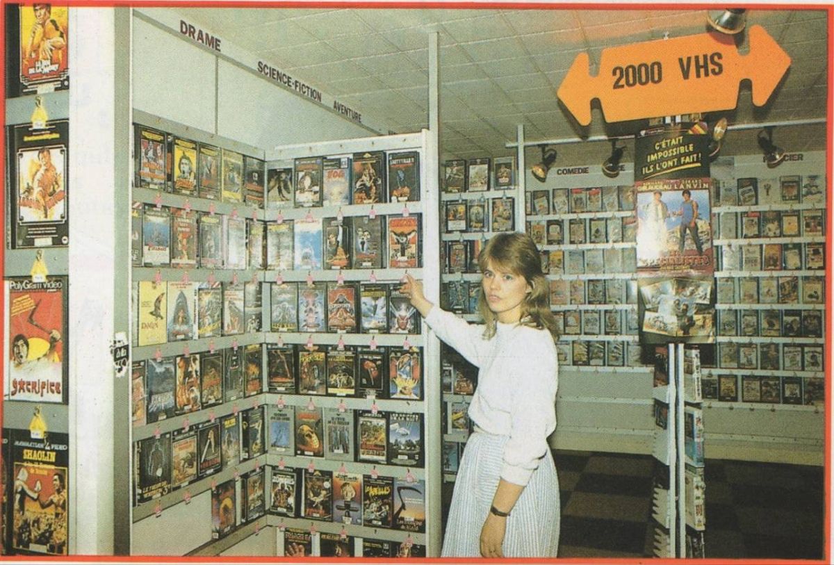 A woman stands in a VHS rental store, reaching toward a shelf filled with movie tapes. Signs above label genres like “Drame” and “Science-Fiction.” A sign in the shape of a tape announces “2000 VHS.”
