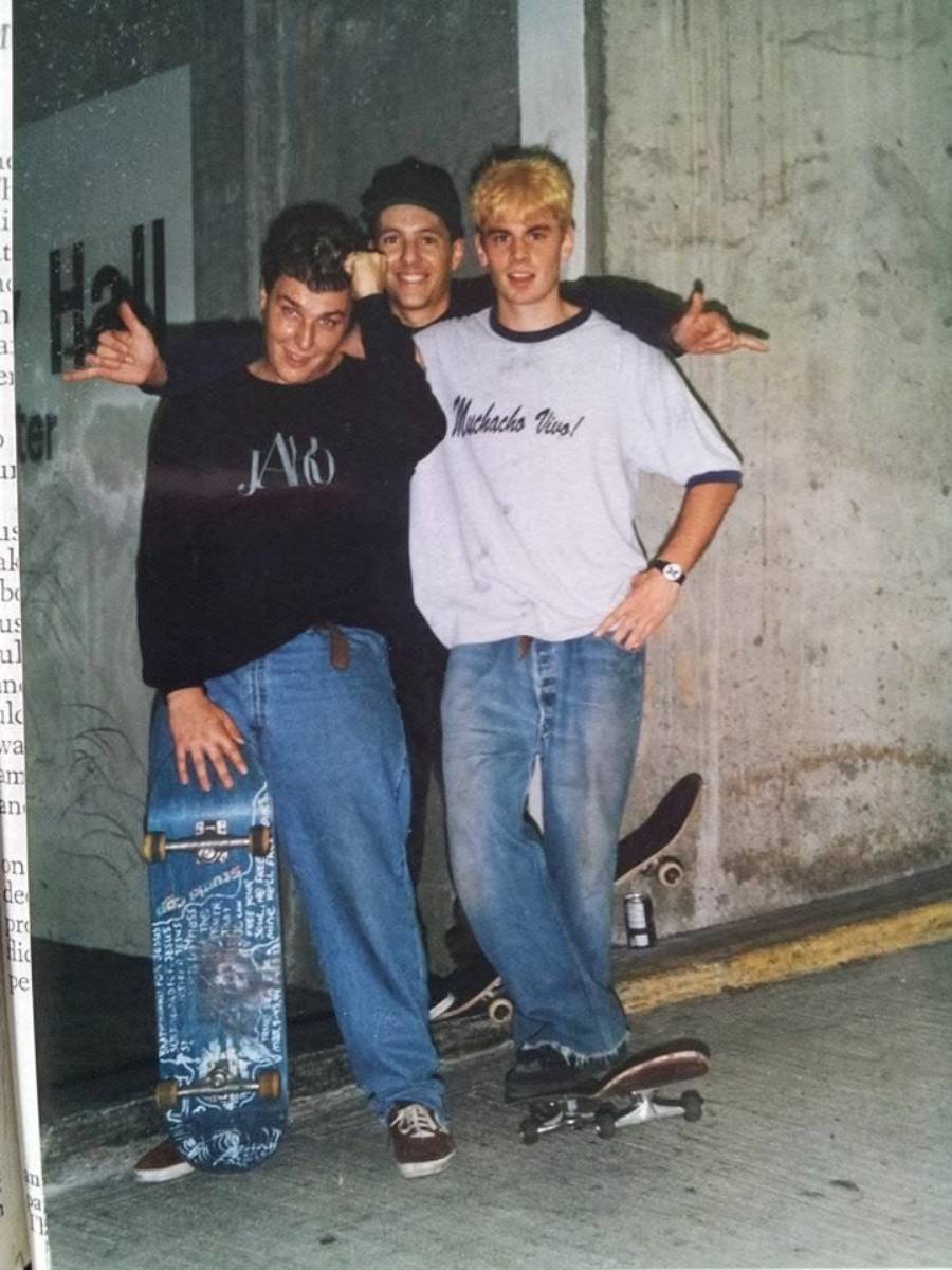 Three young men with skateboards pose and smile in front of a concrete wall. Two stand on the pavement, one stands on a skateboard. They wear casual clothes and look relaxed and happy.