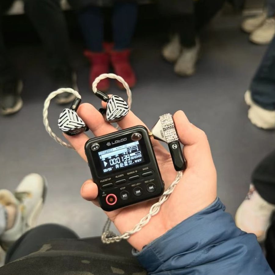 A hand holds a small black music player with braided earphones. People wearing sneakers are seated in the background, suggesting a public transit setting.