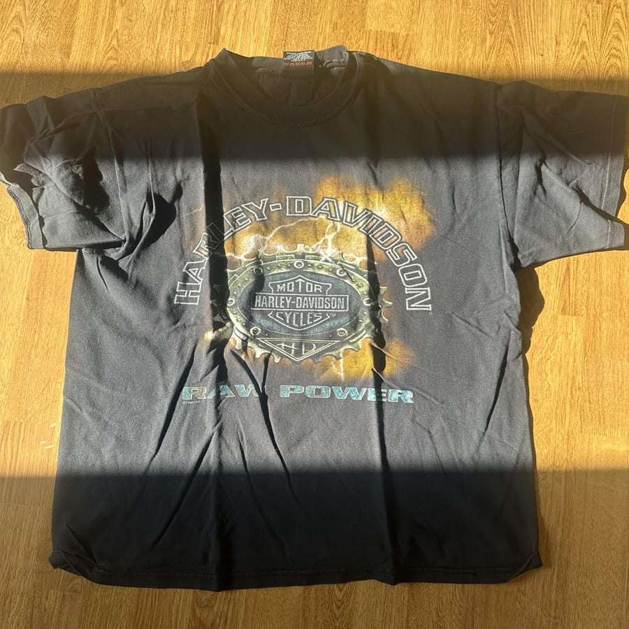A black Harley-Davidson t-shirt with a circular saw blade graphic and the words “Harley-Davidson” and “Raw Power,” shown on a wooden floor with sunlight casting a shadow across the bottom.