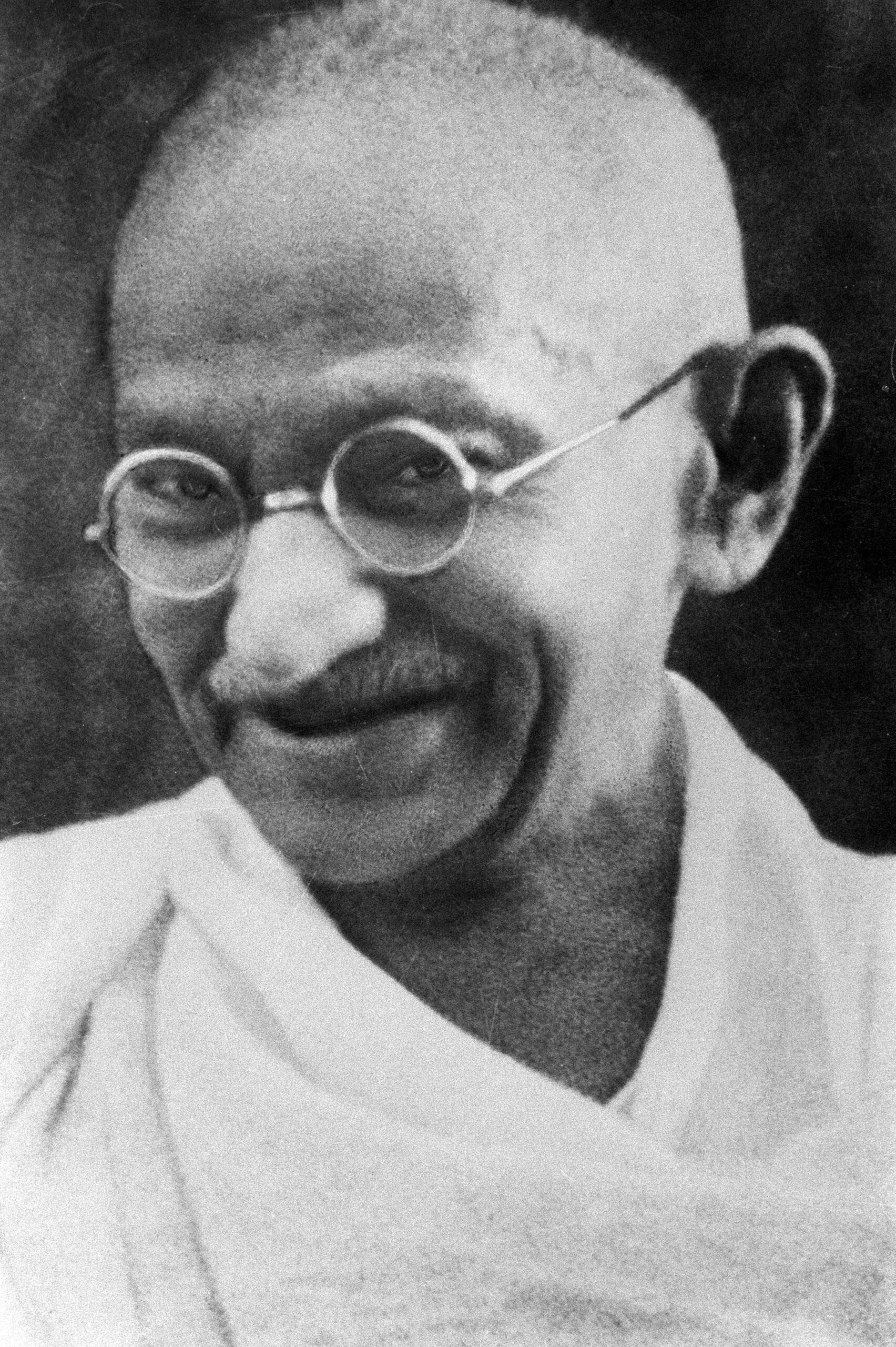 An elderly man with round glasses, a bald head, and a white robe smiles softly, looking slightly to the side. The image is in black and white.