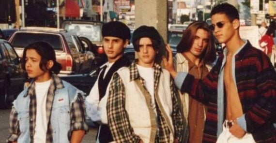 Five teenagers in 1990s streetwear pose on a city sidewalk by a pole, with cars and shops in the background. They wear layered shirts, baggy jeans, and sneakers, exuding a casual, confident attitude.