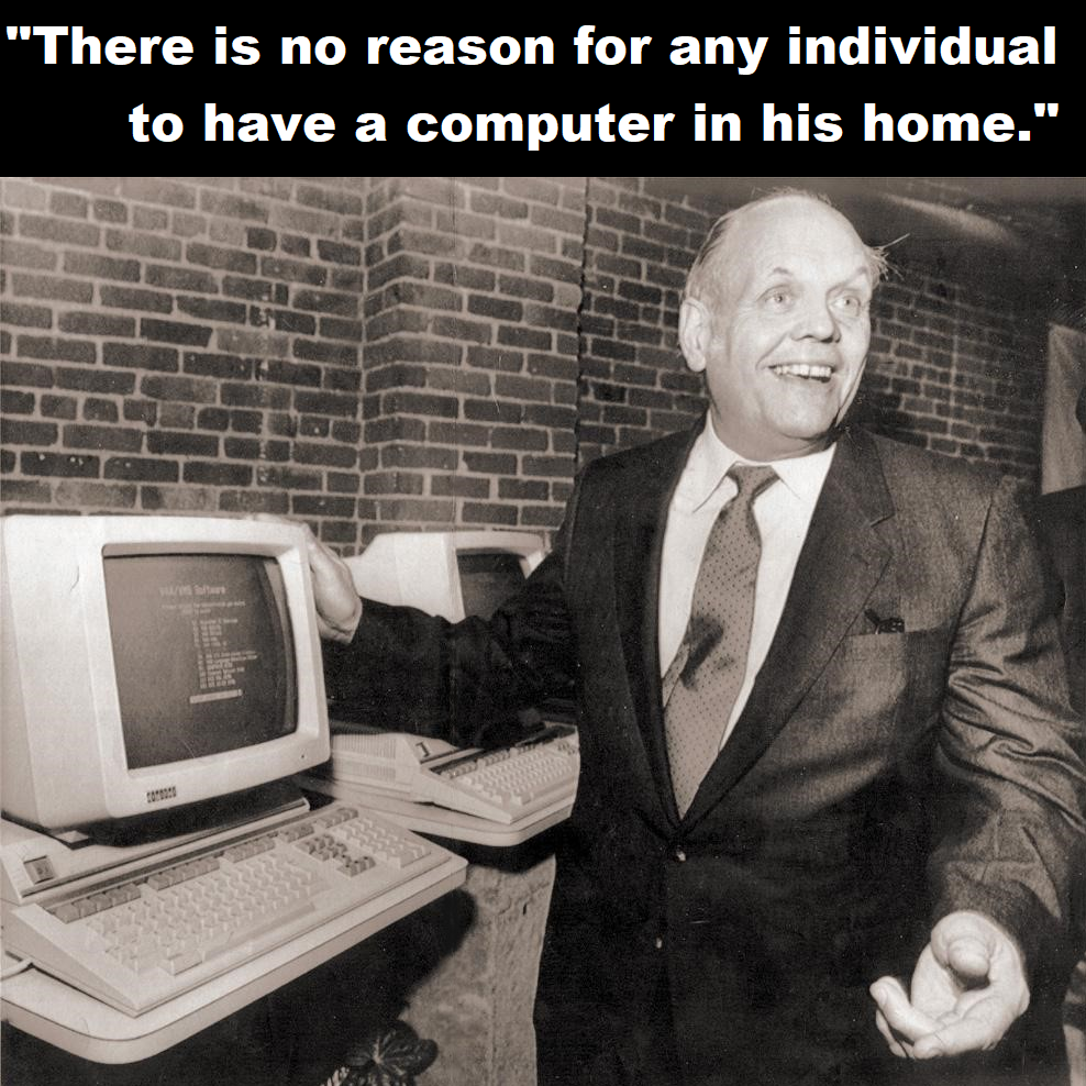 Smiling man in a suit stands by vintage computers. Text above reads, “There is no reason for any individual to have a computer in his home.” Brick wall in the background. Black and white photo.