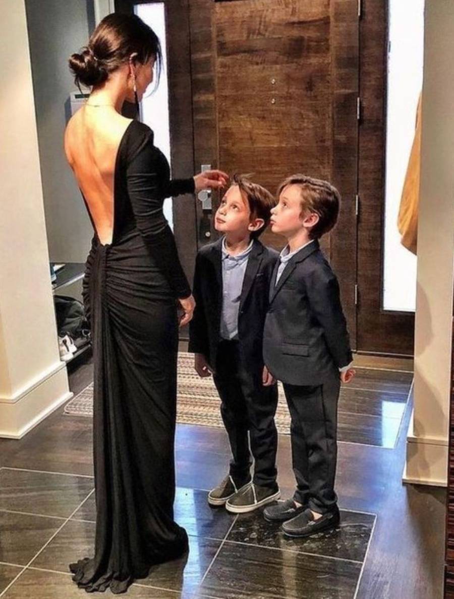 A woman in a backless black dress stands in a hallway, touching the head of one of two young boys in suits who are looking up at her.
