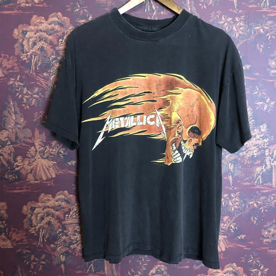 A black Metallica T-shirt hangs on a wooden hanger. The shirt features a fiery, skeletal head graphic and the band’s name in bold letters. The background is a patterned, vintage-style wallpaper.