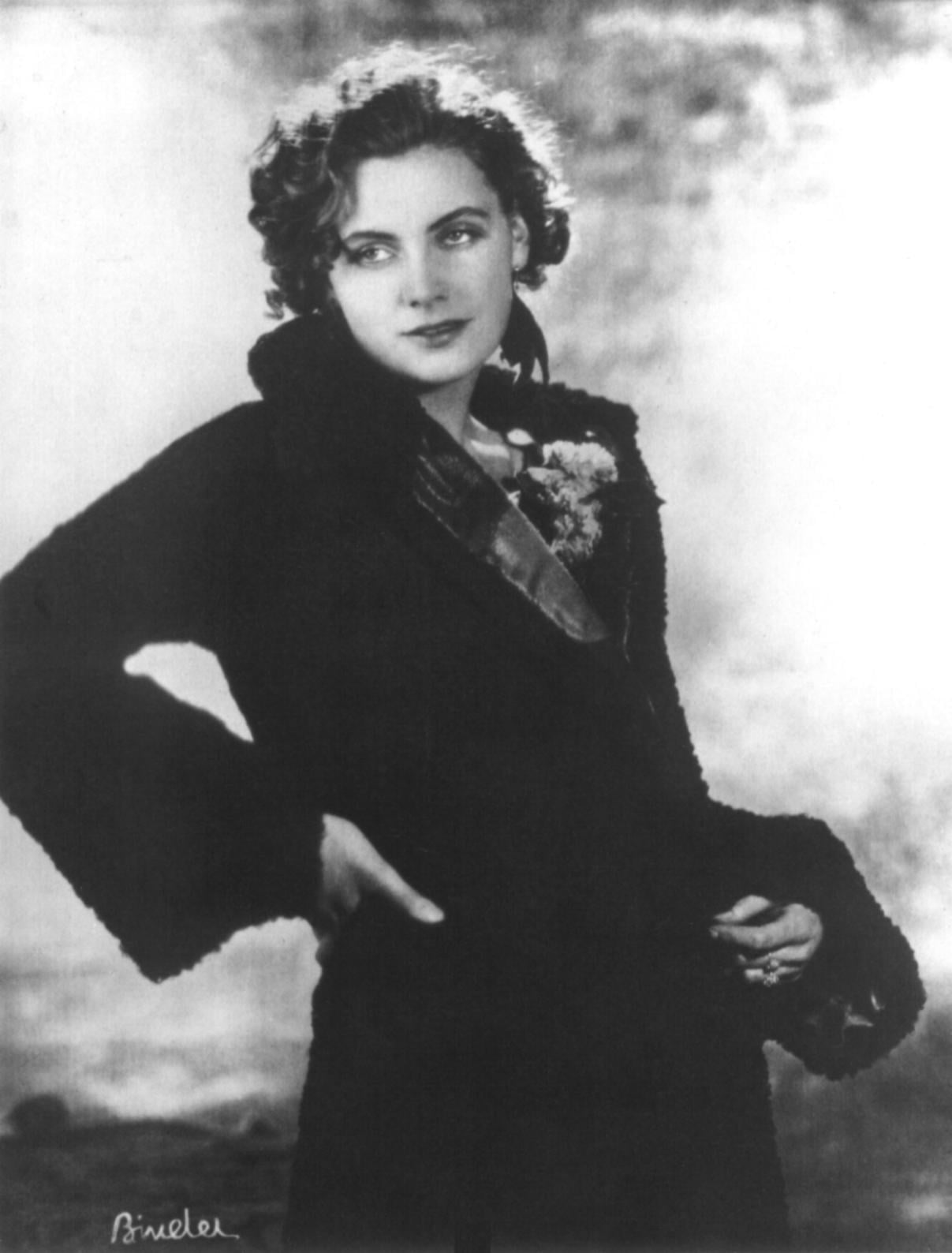 Black-and-white photo of a woman with curly hair wearing a dark coat, looking slightly to the side with one hand on her hip and a soft, contemplative expression.