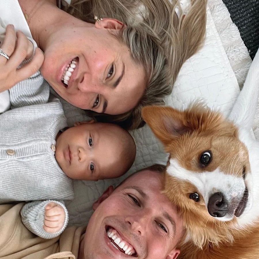 A woman, baby, man, and tan-and-white dog are lying on a white blanket, smiling up at the camera, appearing happy and relaxed together.