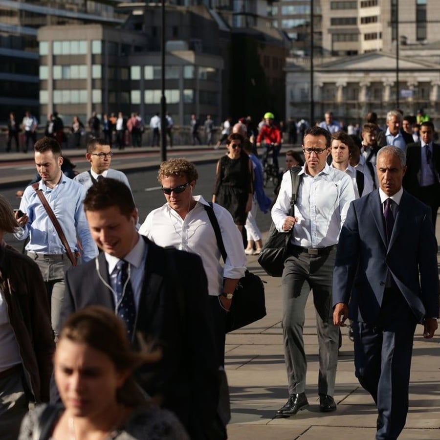 A crowd of people, mostly in business attire, walk on a city sidewalk in sunlight. Some carry bags and wear sunglasses. Tall buildings are visible in the background.