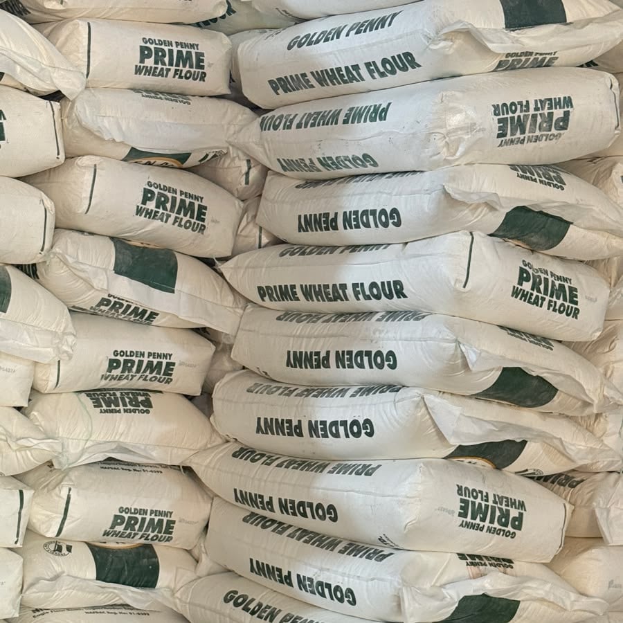 A large stack of white sacks labeled “Golden Penny Prime Wheat Flour” arranged in rows, with some bags oriented upside down and sideways, creating a dense, textured pattern.