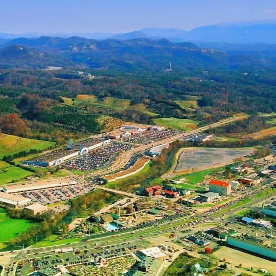 Aerial view of a busy town with parking lots, roads, and buildings surrounded by green hills and mountains in the distance under a clear blue sky.
