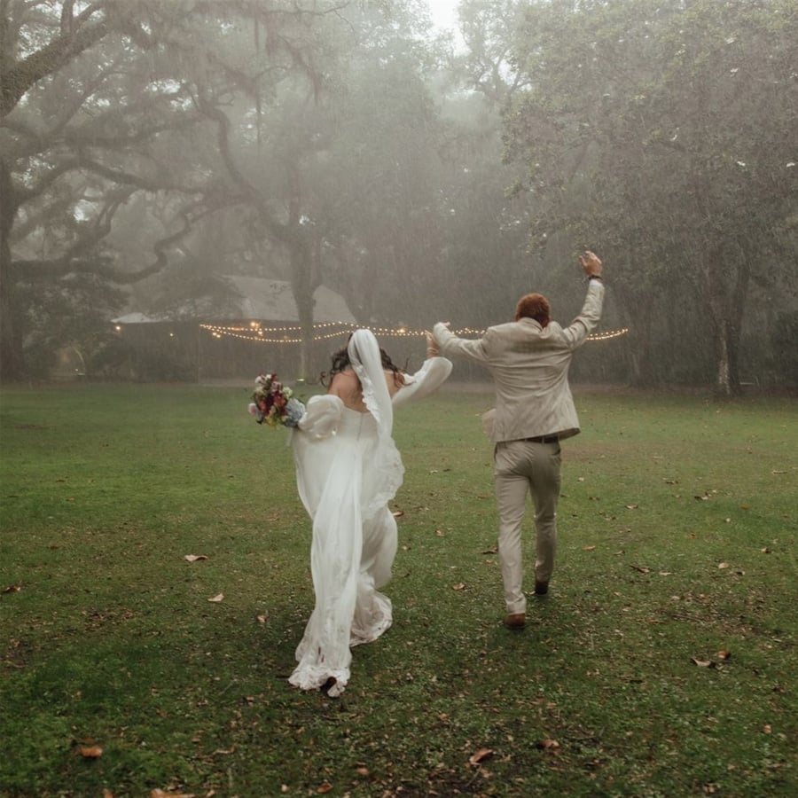 A bride and groom joyfully run through a grassy field in the rain, holding hands. The bride holds a bouquet, and string lights glow in the background among large trees.