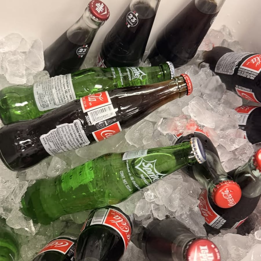 Glass bottles of Coca-Cola and Sprite are lying on a bed of ice, with condensation visible on the bottles, keeping the drinks cold.
