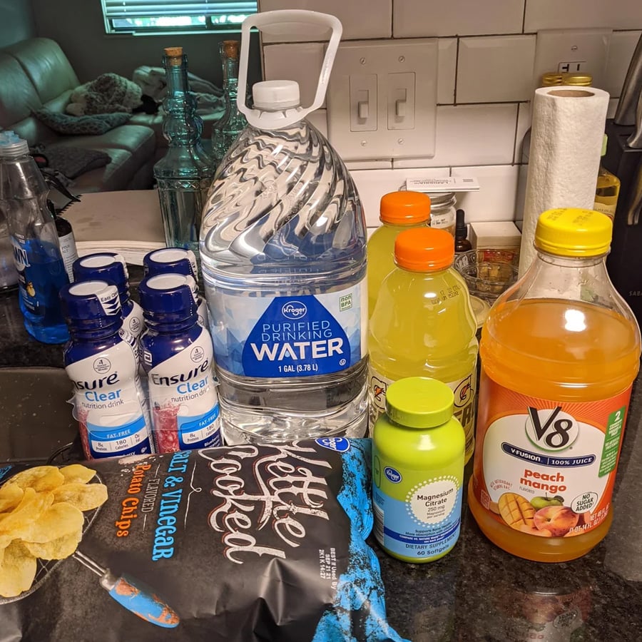 A variety of drinks, chips, and snacks on a kitchen counter, including bottled water, Ensure Clear, V8 juice, lemonade, a green bottle, and a bag of salt & vinegar kettle chips.