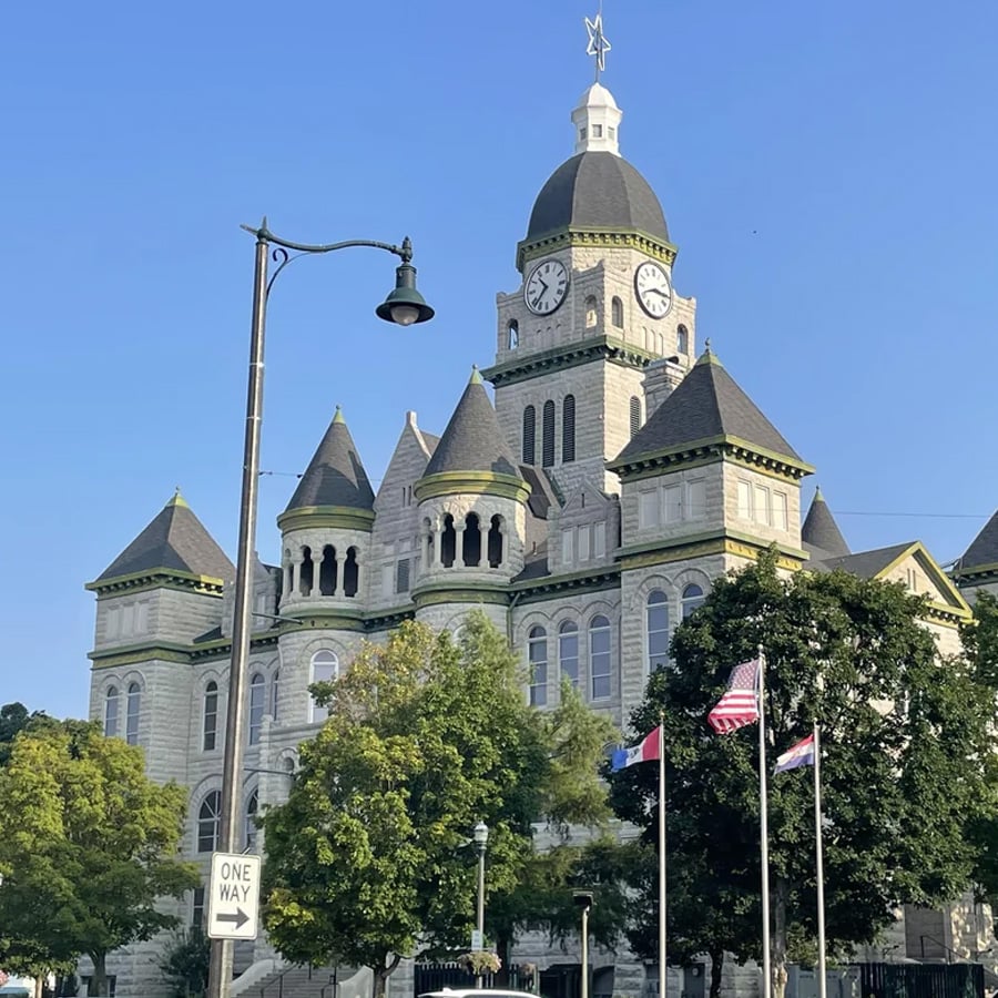 A historic stone courthouse with a clock tower, turrets, and a star on top stands behind trees and flags under a clear blue sky. A one-way street sign and a streetlamp are in the foreground.