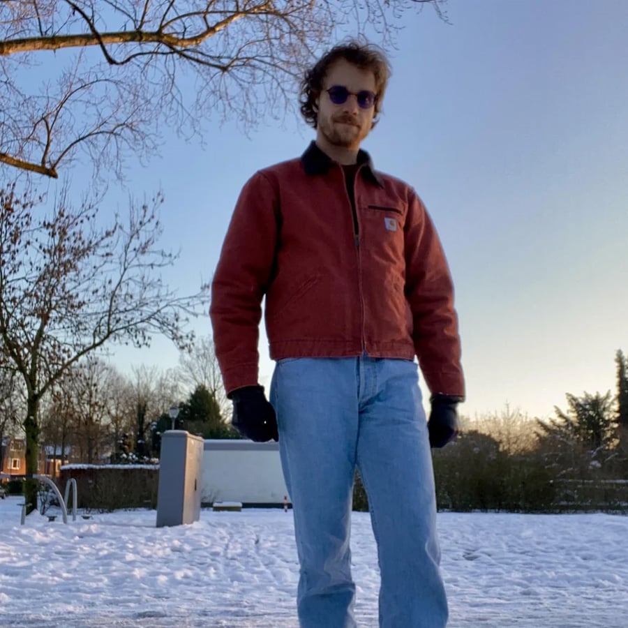 A person in a red jacket, blue jeans, gloves, and sunglasses stands on snow-covered ground with bare trees and a clear sky in the background.
