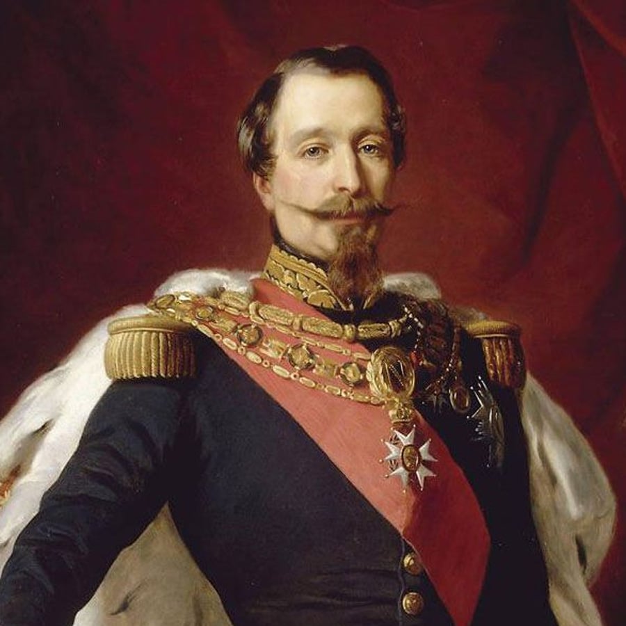 A man in a formal military uniform with gold decorations, a red sash, and a white fur cape stands against a dark red background. He has a mustache, goatee, and short brown hair.