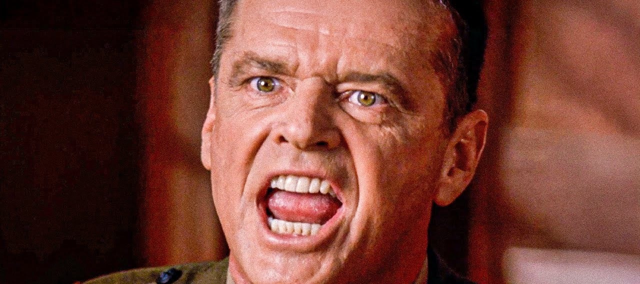 A man in uniform with an intense expression is shouting, his mouth wide open and teeth visible. The background is blurred, focusing attention on his face and emotion.