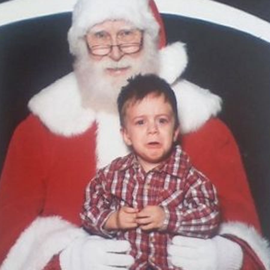 A child in a plaid shirt looks upset while sitting on Santa Claus’s lap. Santa is smiling and wearing glasses, a red suit, and a white beard against a dark background.