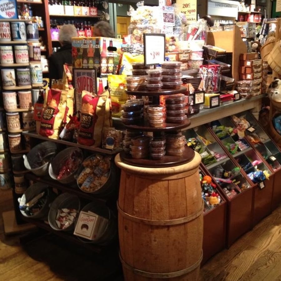 A display in a general store shows snacks, candy, and baked goods on shelves and a barrel, with chips, tins, jars, and bins of colorful sweets arranged in a cozy, rustic setting.
