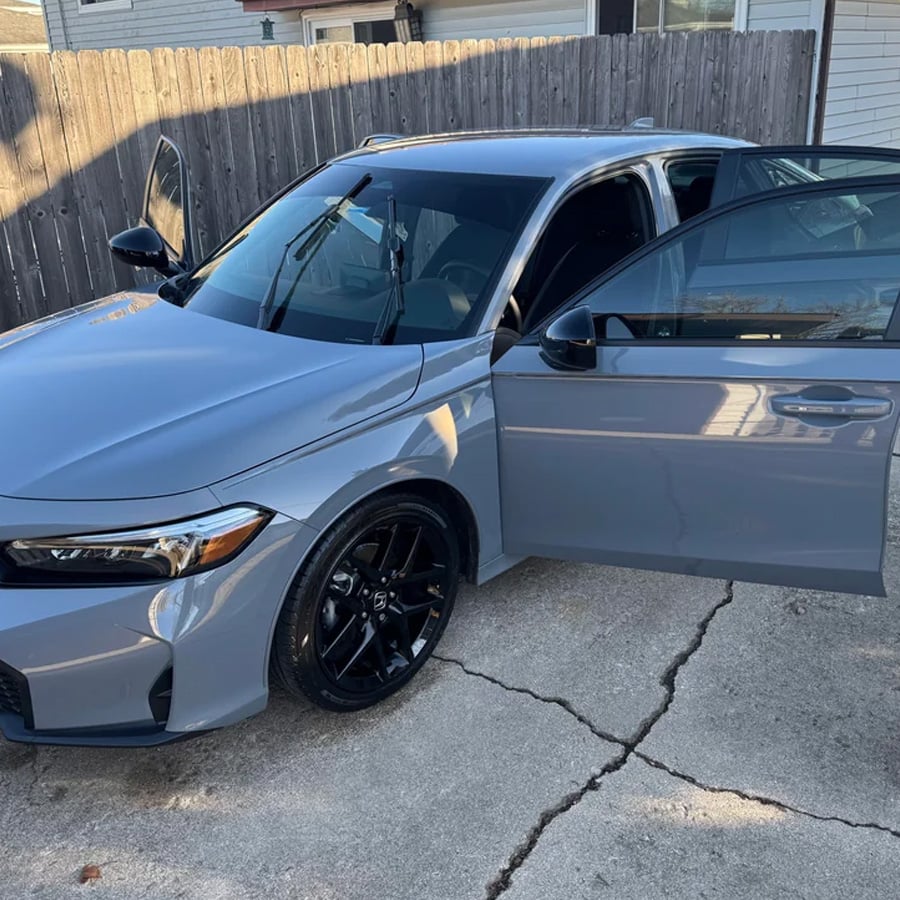 A gray sedan with black wheels is parked on a driveway. Both front doors are open. A wooden fence and a house are visible in the background.