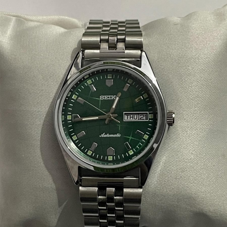 A stainless steel Seiko watch with a green face, silver hour markers, and a day-date window showing “THU 21,” displayed on a white cushioned surface.