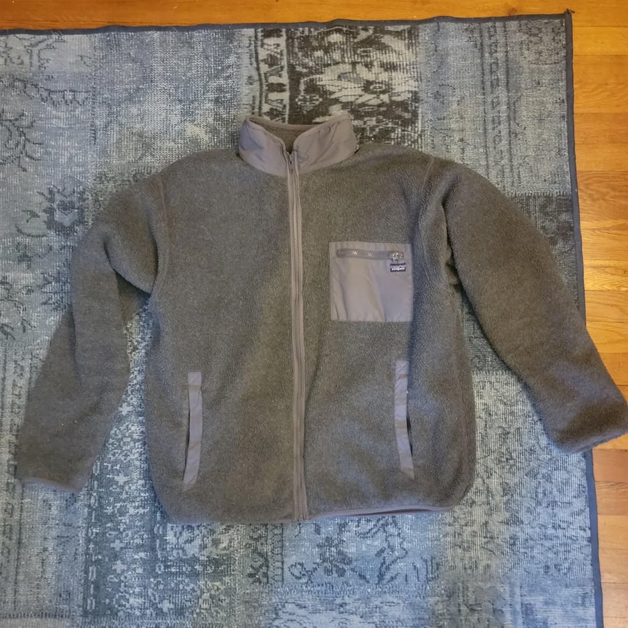 A gray fleece zip-up jacket with a stand-up collar and a chest pocket is laid flat on a blue patterned rug with wooden floor visible at the top.