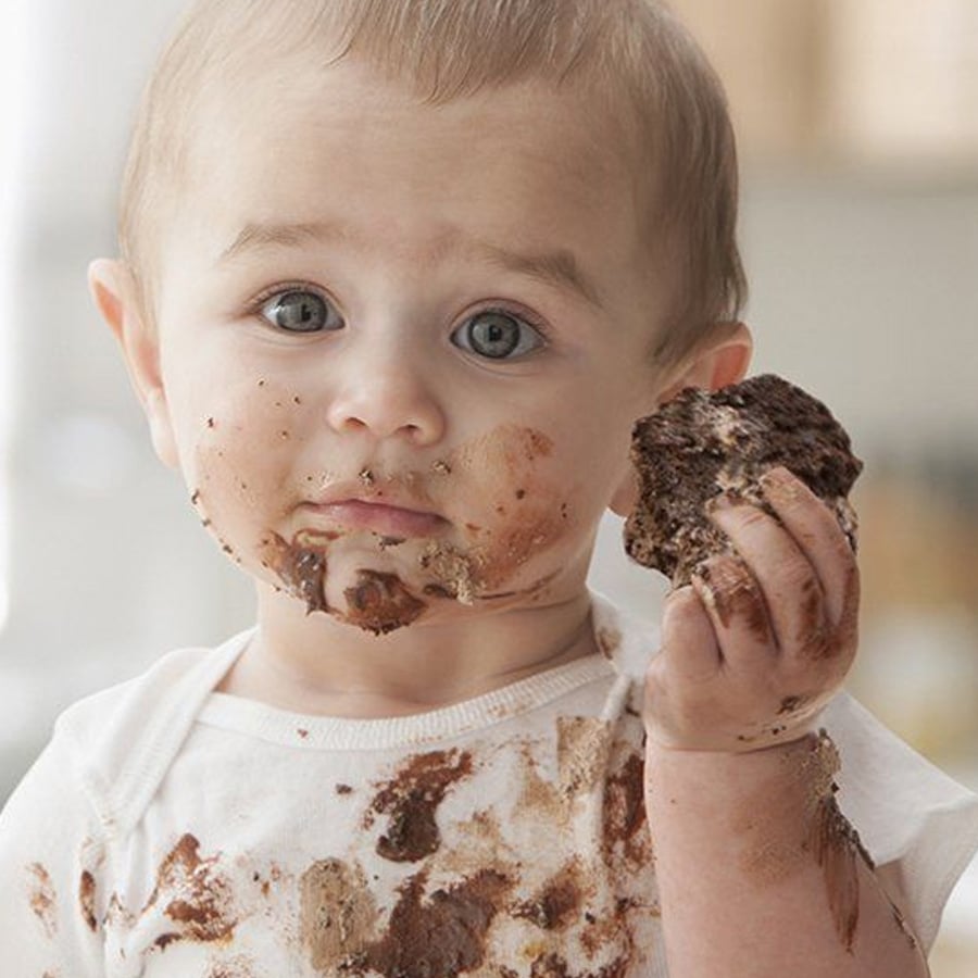 A baby with blue eyes and light hair is holding a chocolate-covered cookie, with chocolate smeared on their face, hands, and white shirt.
