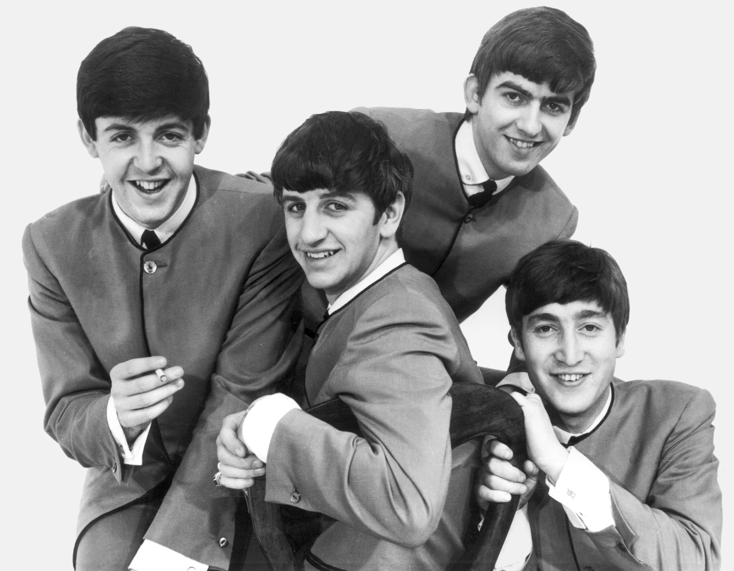 Four young men in matching suits pose together, smiling and leaning closely. The photo is black and white with a plain background.