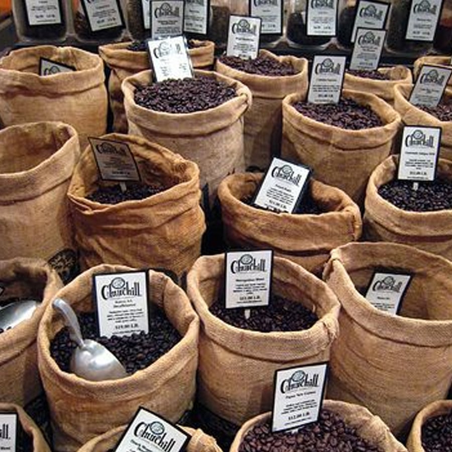 Several burlap sacks filled with different types of coffee beans, each labeled with a Churchill Coffee sign, are displayed together. A metal scoop rests in one of the sacks.