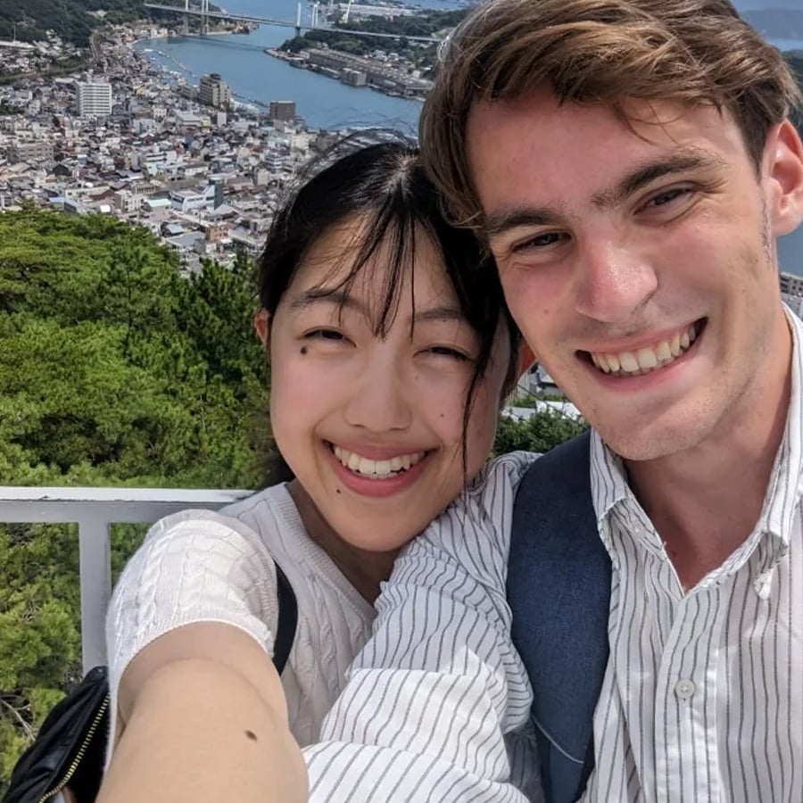 Two people smiling and taking a selfie together outdoors, with a scenic city and river view in the background.