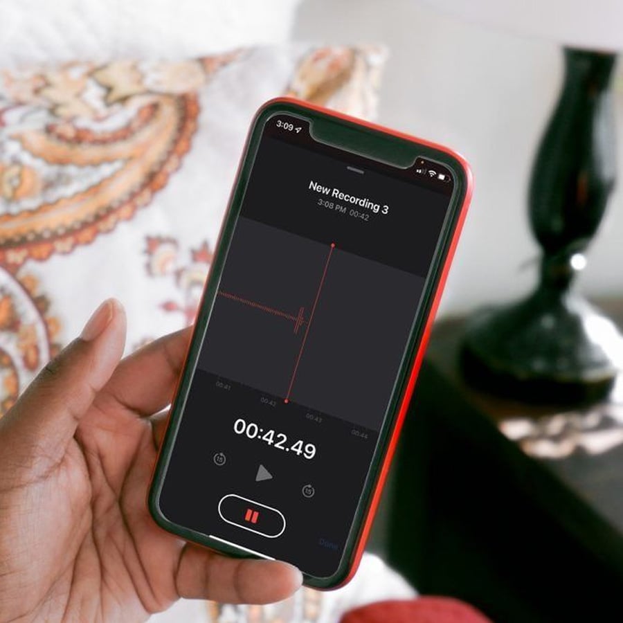 A hand holds a smartphone displaying a voice recording app with a 42-second recording in progress. The background shows a patterned pillow and part of a lamp.
