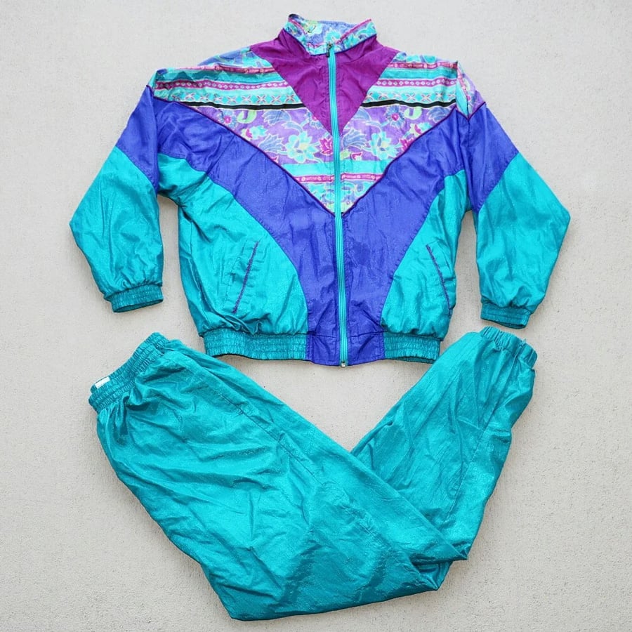 A vintage turquoise, purple, and magenta tracksuit with a floral pattern on the jacket, displayed flat on a light beige surface.