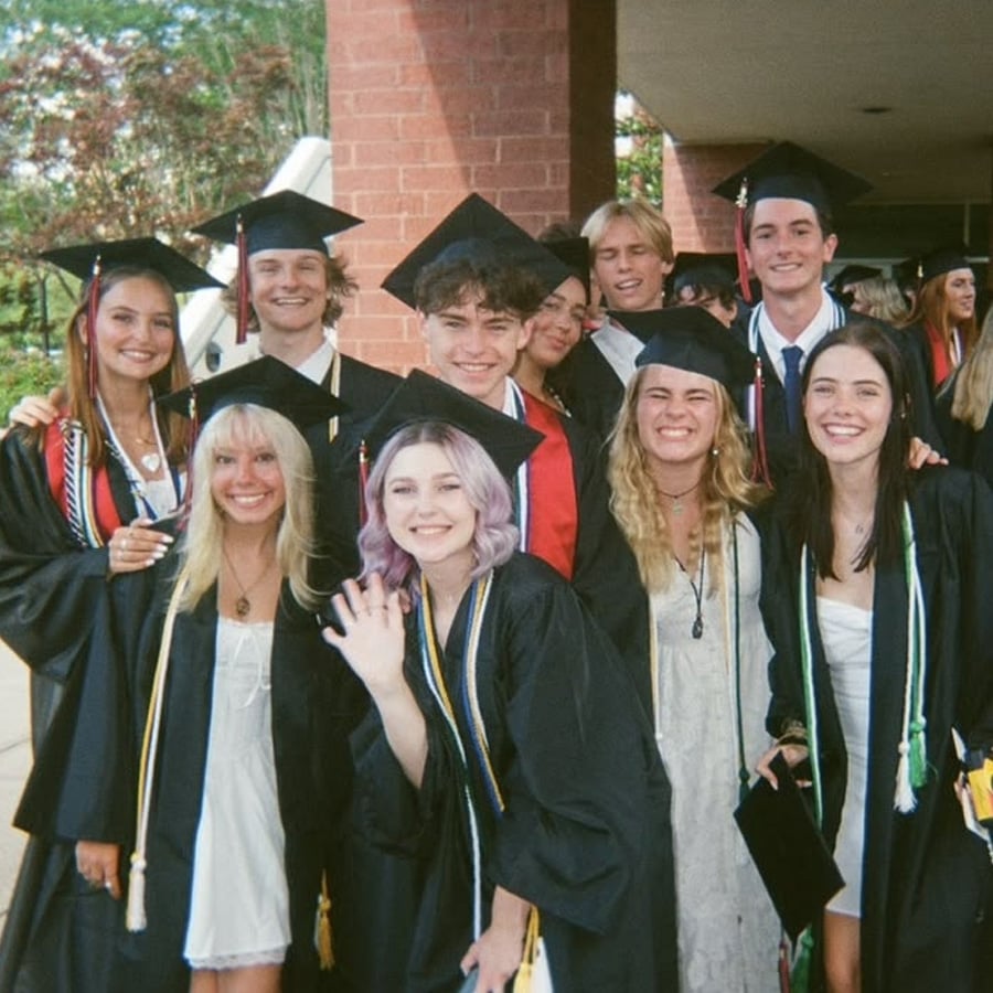 A group of graduates in caps and gowns smile and pose together outdoors, some holding diplomas and wearing honor cords, with trees and a building in the background.